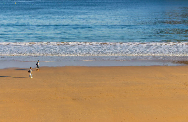 San Sebastian, Spanyolország - 10 / 11 / 2019: Ismeretlen emberek sétálnak a tengerparton. Turisták a Vizcayai-öbölben San Sebastianban ősszel. Széles antenna strand a reggeli napsütésben. Egészséges és nyugodt életmód. Aktív személyek. - Fotó, kép