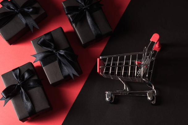 Black Friday sale shopping concept, Gift box wrapped black paper and black bow ribbon present and the shopping cart, studio shot isolated on red and dark background - Fotografie, Obrázek