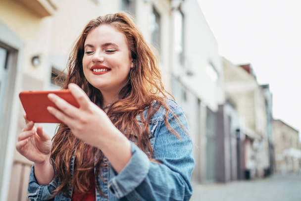 Smiling red curly long hair caucasian teen girl walking on the street and browsing the internet using the modern smartphone. Modern people with technology devices concept image. - Фото, зображення
