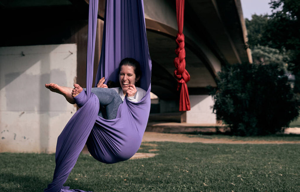 A happy Caucasian woman using her aerial stretch fabric as a hammock while practicing in a park - Foto, Bild