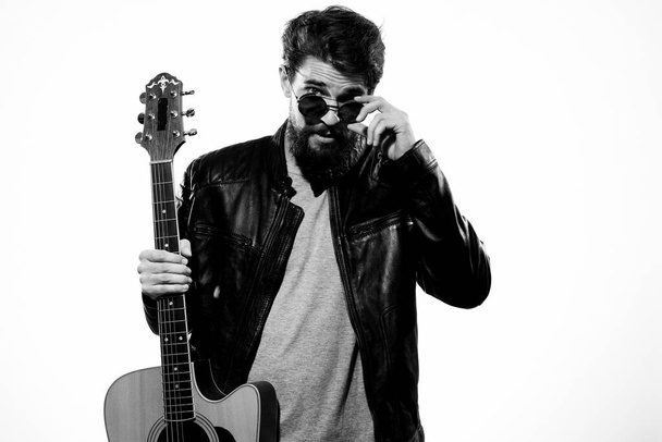 Un hombre sostiene una guitarra en sus manos chaqueta de cuero negro gafas oscuras música rendimiento fondo claro - Foto, Imagen