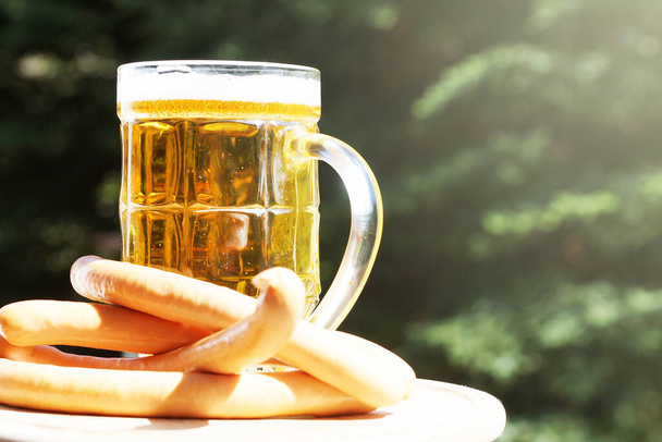стеклянная кружка с пивом и сосисками на фоне природы в солнечный день - Фото, изображение