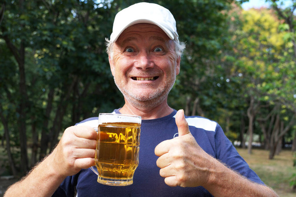 смеющийся человек держит кружку пива в одной руке, с другой стороны показывает, что в парке все в порядке - Фото, изображение