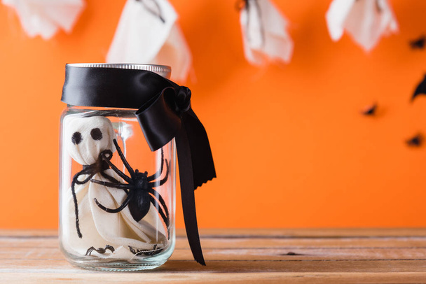 Funny Halloween day decoration party, Baby white ghost crafts scary face in jar glass on wood table, studio shot isolated on orange background have spider and bats, Happy holiday DIY concept - Foto, Bild