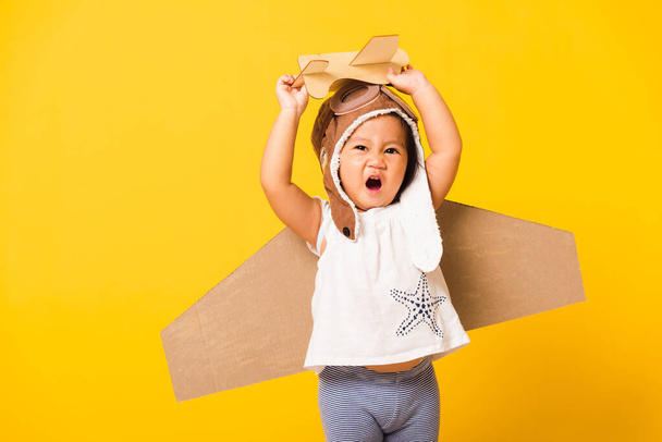 Happy Asian beautiful funny baby little girl smile wear pilot hat play and goggles with toy cardboard airplane wings fly hold plane toy, studio shot isolated yellow background, Startup freedom concept - Фото, изображение