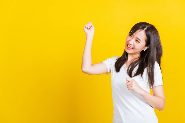 Portrait Asian of a happy beautiful young woman wear wireless earphone listening to music from smartphone having fun and dancing to sounds of favorite music studio shot isolated on yellow background - 写真・画像