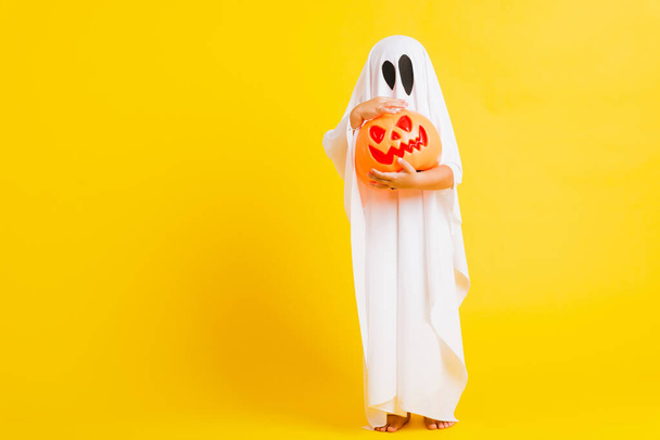 Funny Halloween Kid Concept, little cute child with white dressed costume halloween ghost scary he holding orange pumpkin ghost on hand, studio shot yellow on white background - Photo, image