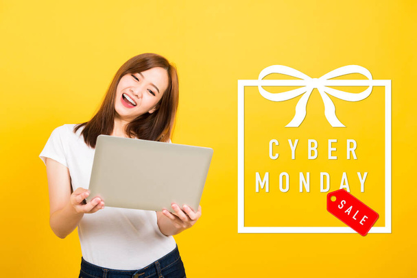 Asian happy portrait beautiful cute young woman teen smile standing hold laptop computer and excited celebrating success with Cyber Monday in the gift box on space, studio shot yellow background - Photo, image