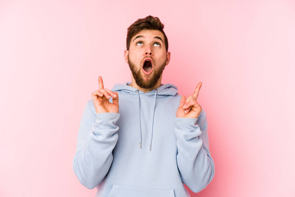 Young caucasian man isolated on pink background pointing upside with opened mouth. - Фото, изображение