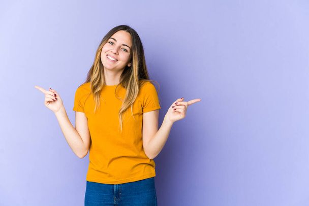 Young caucasian woman isolated on purple background pointing to different copy spaces, choosing one of them, showing with finger. - Photo, Image