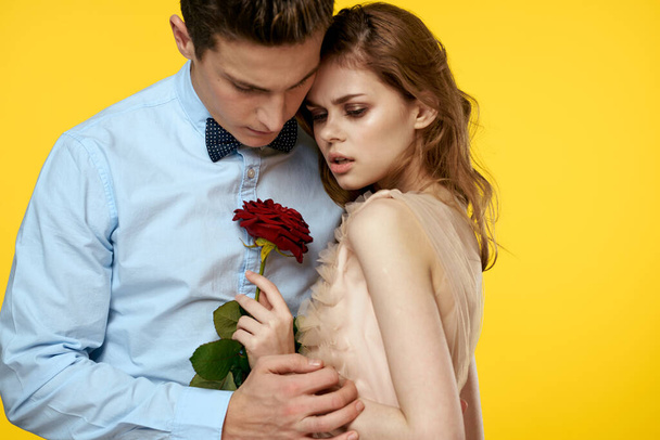 Ένα ερωτευμένο νεαρό ζευγάρι με ένα κόκκινο τριαντάφυλλο στα χέρια αγκαλιάζονται σε κίτρινο φόντο. - Φωτογραφία, εικόνα