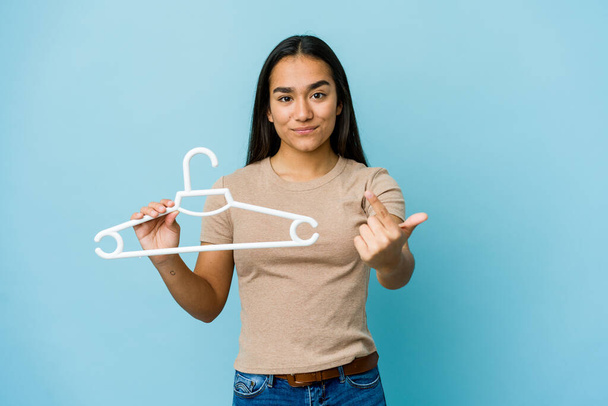 Young asian woman holding a hanger isolated on blue background pointing with finger at you as if inviting come closer. - Photo, Image