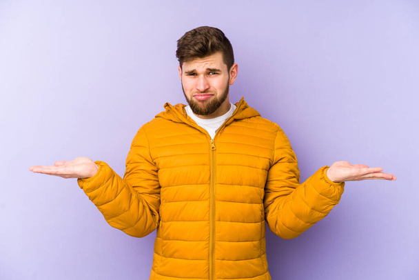 Young man isolated on purple background confused and doubtful shrugging shoulders to hold a copy space. - Photo, Image