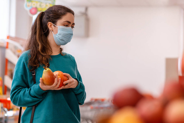 Ψώνια. Μια νεαρή λευκή γυναίκα με μια ιατρική μάσκα στο πρόσωπό της, κρατώντας ένα αχλάδι και ένα ρόδι, κοιτάζοντας ένα παράθυρο με φρούτα. Αντιγραφή χώρου. Η έννοια της αγοράς φρούτων και την προστασία από coronovirus. - Φωτογραφία, εικόνα