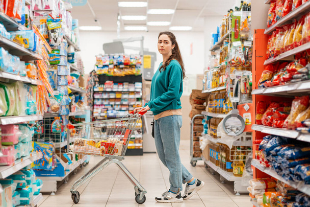 Una joven guapa caucásica vestida con ropa casual posa con un carrito de comestibles en el pasillo de una tienda. El concepto de comprar y comprar alimentos. - Foto, imagen