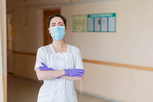 Coronavirus. Un medico in camice da laboratorio, maschera medica e guanti posa in un corridoio d'ospedale con le braccia incrociate sul petto. Il concetto di medici eroi e la lotta contro la pandemia di coronavirus. - Foto, immagini
