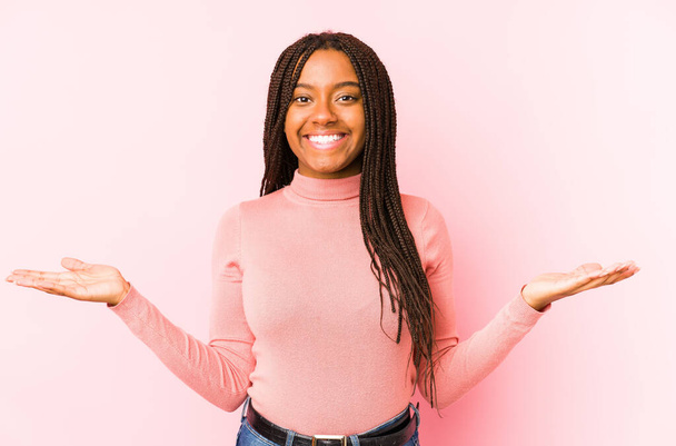 Νεαρή Αφροαμερικανή, απομονωμένη σε ροζ φόντο, κάνει κλίμακα με τα χέρια, αισθάνεται ευτυχισμένη και σίγουρη.. - Φωτογραφία, εικόνα