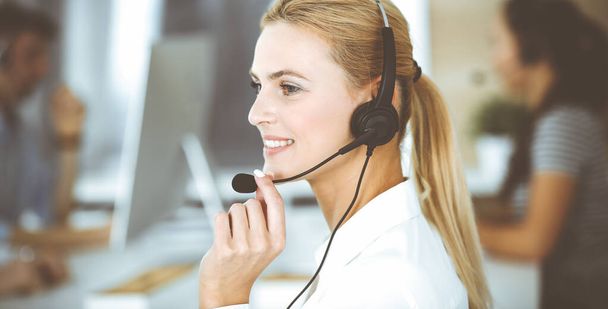 Blonde business woman using headset for communication and consulting people at customer service office. Call center. Group of operators at work - Photo, image