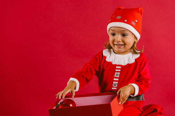 Adorable little girl in a christmas costume with surprised opens a new year gift on a red background with blank space - Valokuva, kuva