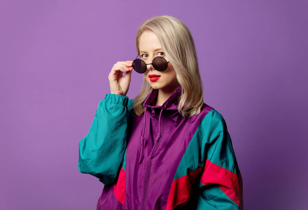 Стильная блондинка в 80-х ветровка и шумные солнечные очки на фиолетовом фоне  - Фото, изображение