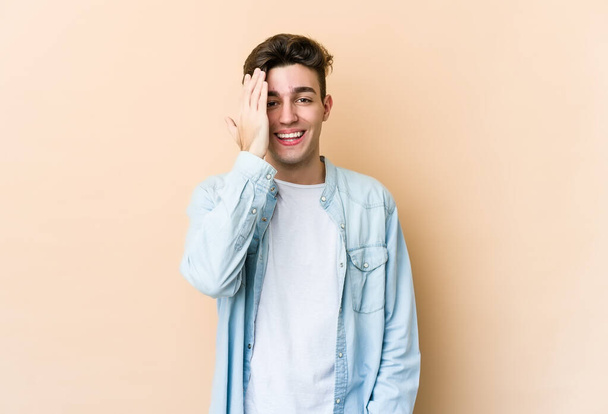 Young caucasian man isolated on beige background having fun covering half of face with palm. - Photo, image