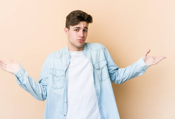 Young caucasian man isolated on beige background doubting and shrugging shoulders in questioning gesture. - Photo, Image