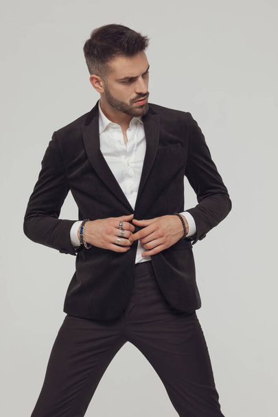 Confident fashion unbuttoning his jacket while standing with legs spread on gray studio background - Foto, afbeelding