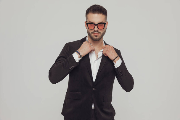 Serious fashion model fixing collar and wearing sunglasses while standing on gray studio background - Photo, image