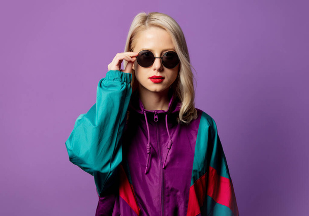 Style blonde in 80s windbreaker and roud sunglasses on purple background  - Фото, изображение