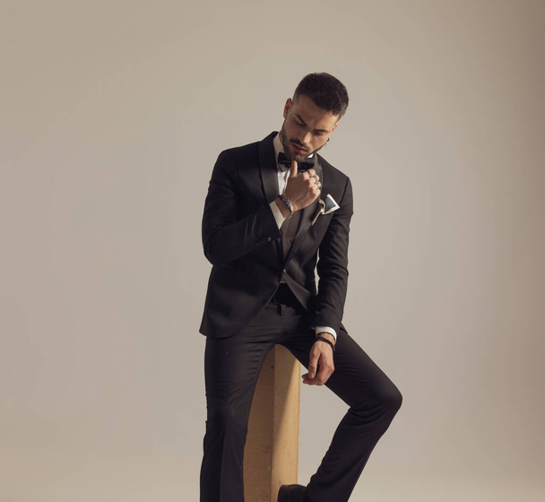 Thoughtful groom holding hand on chin and looking down, wearing tuxedo while sitting on gray studio background - Photo, Image
