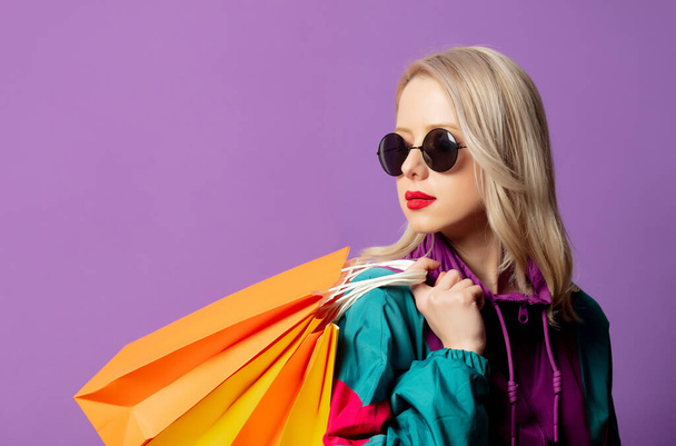 Stylische Frau in 80er-Jahre-Windjacke und Sonnenbrille hält Einkaufstüten auf lila Hintergrund - Foto, Bild