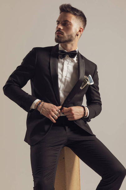 Confident groom unbuttoning his jacket and looking away, wearing tuxedo while sitting on gray studio background - Fotografie, Obrázek