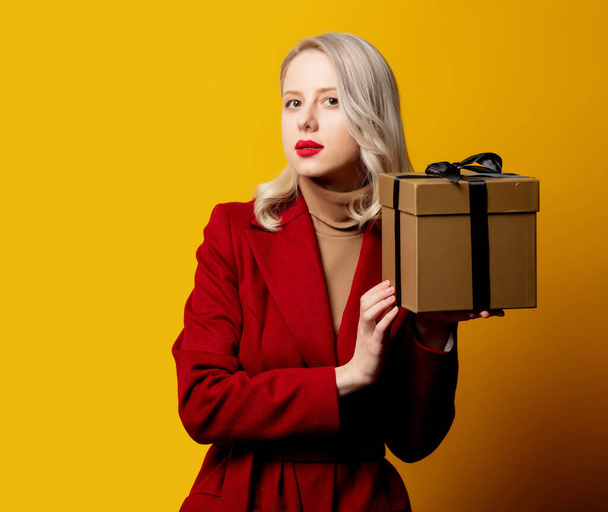 Mujer rubia sorprendida en abrigo rojo con caja de regalo sobre fondo amarillo  - Foto, Imagen