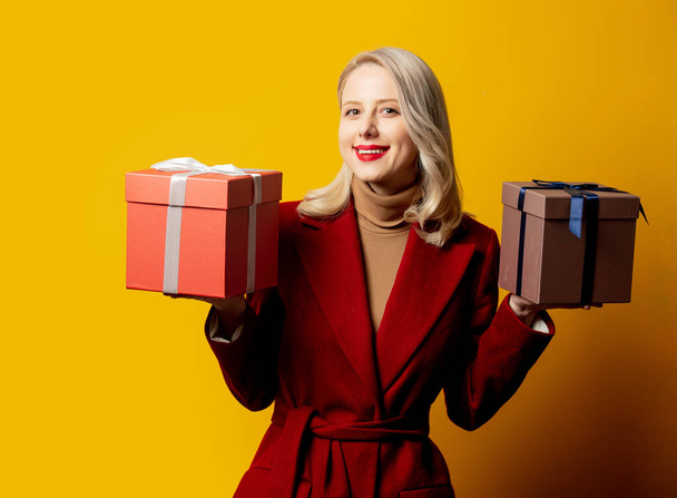 Blonde smiling woman in red coat with gift boxes on yellow background - Fotó, kép