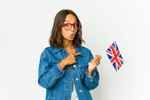 Jovem mulher latina segurando uma bandeira inglesa isolada no fundo branco sorrindo e apontando para o lado, mostrando algo no espaço em branco. - Foto, Imagem