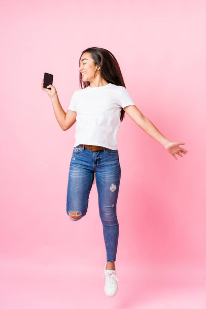 Young indian woman holding phone jumping on pink background - Fotografie, Obrázek
