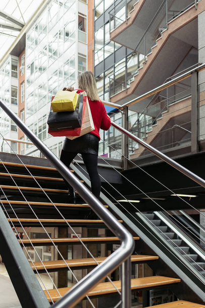 Ενήλικες γυναίκα φορώντας ένα κόκκινο πουλόβερ με τα πόδια μέχρι τις σκάλες ενός εμπορικού κέντρου μεταφέρουν πολύχρωμες τσάντες ψώνια. - Φωτογραφία, εικόνα