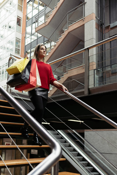 Aikuinen nainen yllään punainen villapaita kävelee alas portaita ostoskeskuksen kuljettaa värikkäitä ostoskasseja. - Valokuva, kuva