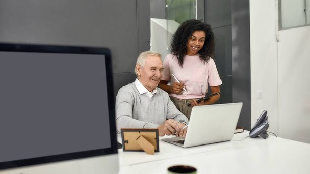 Aged man, senior intern looking at laptop while showing results to his young colleague, Friendly female worker mentoring and training new employee, monitoring his progress at work - Foto, imagen