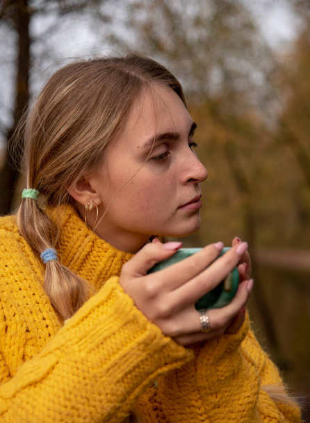 Portrait of a blonde girl in a yellow knitted sweater with a green Cup in her hands on a blurred natural background. - Φωτογραφία, εικόνα
