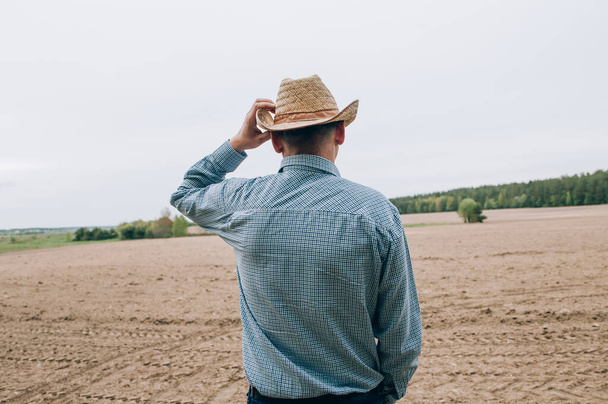 Man farmer agronomist in jeans and shirt stands back in field after haymaking, with tablet looking into the distance. Rural business, agricultural industry, freedom after work, concept - Photo, Image