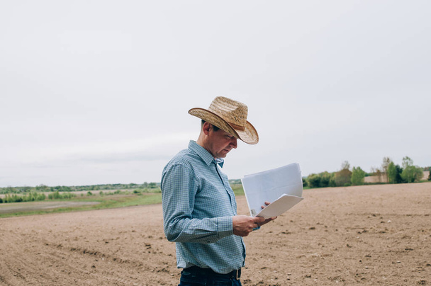Man farmer agronomist in jeans and shirt stands back in field after haymaking, with tablet looking into the distance. Rural business, agricultural industry, freedom after work, concept - Фото, изображение