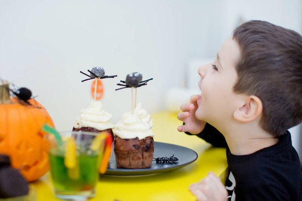 Le garçon mange des bonbons à la fête d'Halloween. Enfant caucasien garçon mange muffins Halloween ou cupcakes avec crème blanche et des araignées décorations. Nourriture savoureuse et effrayante pendant la fête d'Halloween - Photo, image