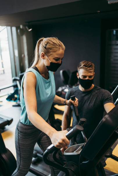 Young fit and attractive woman at body workout in modern gym together with her personal fitness instructor or coach. They keeping distance and wearing protective face masks. Coronavirus world pandemic and sport theme. - Foto, Imagem