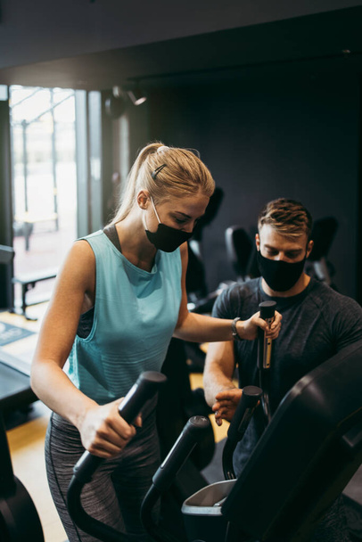 Young fit and attractive woman at body workout in modern gym together with her personal fitness instructor or coach. They keeping distance and wearing protective face masks. Coronavirus world pandemic and sport theme. - Fotoğraf, Görsel