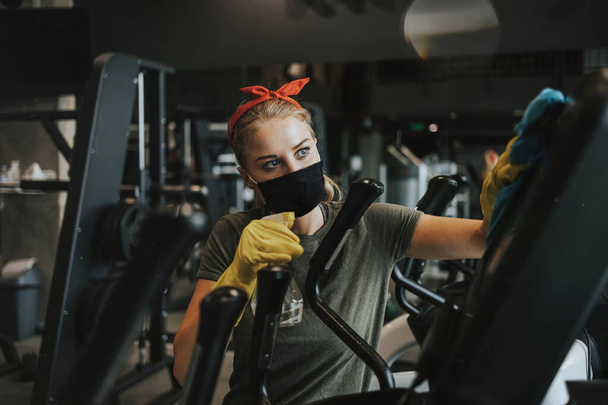 Young female worker disinfecting cleaning and weeping expensive fitness gym equipment with alcohol sprayer and cloth. Coronavirus global world pandemic and health protection safety measures. - Photo, image