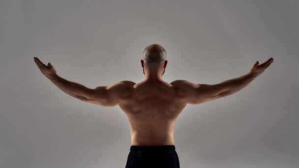 Perfect shape. Rear view of muscular strong man with naked torso standing with outstretched arms isolated over grey background - Zdjęcie, obraz