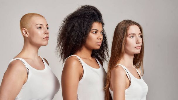 Portrait of three confident young diverse women wearing white shirts looking aside while posing together isolated over grey background - Φωτογραφία, εικόνα