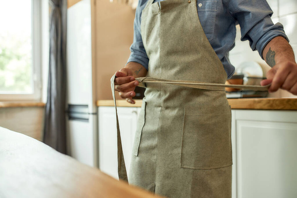 Cropped shot of young man, professional cook tying his apron while getting ready to prepare a meal, standing in the kitchen. Cooking at home concept - 写真・画像