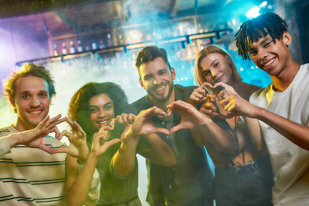 ナイトクラブの時間だ。カメラの前で一緒にポーズをとりながらハートサインを見せる若い男女。バーでパーティーに遊びに来る友達の多人種グループ - 写真・画像